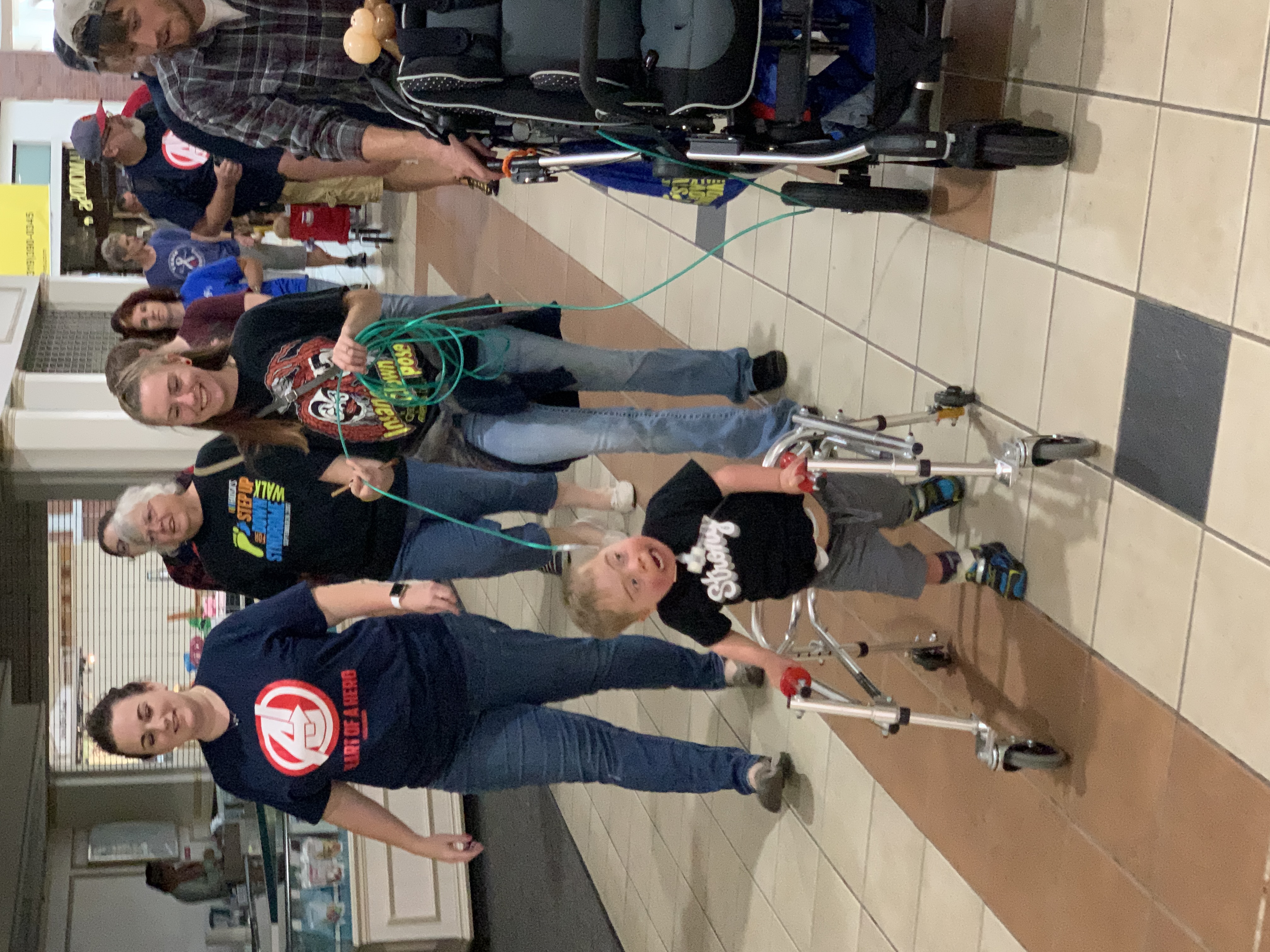 Boy with Down syndrome wearing afo and smo using a reverse walker  in  a shopping mall wearing a shirt that says strong