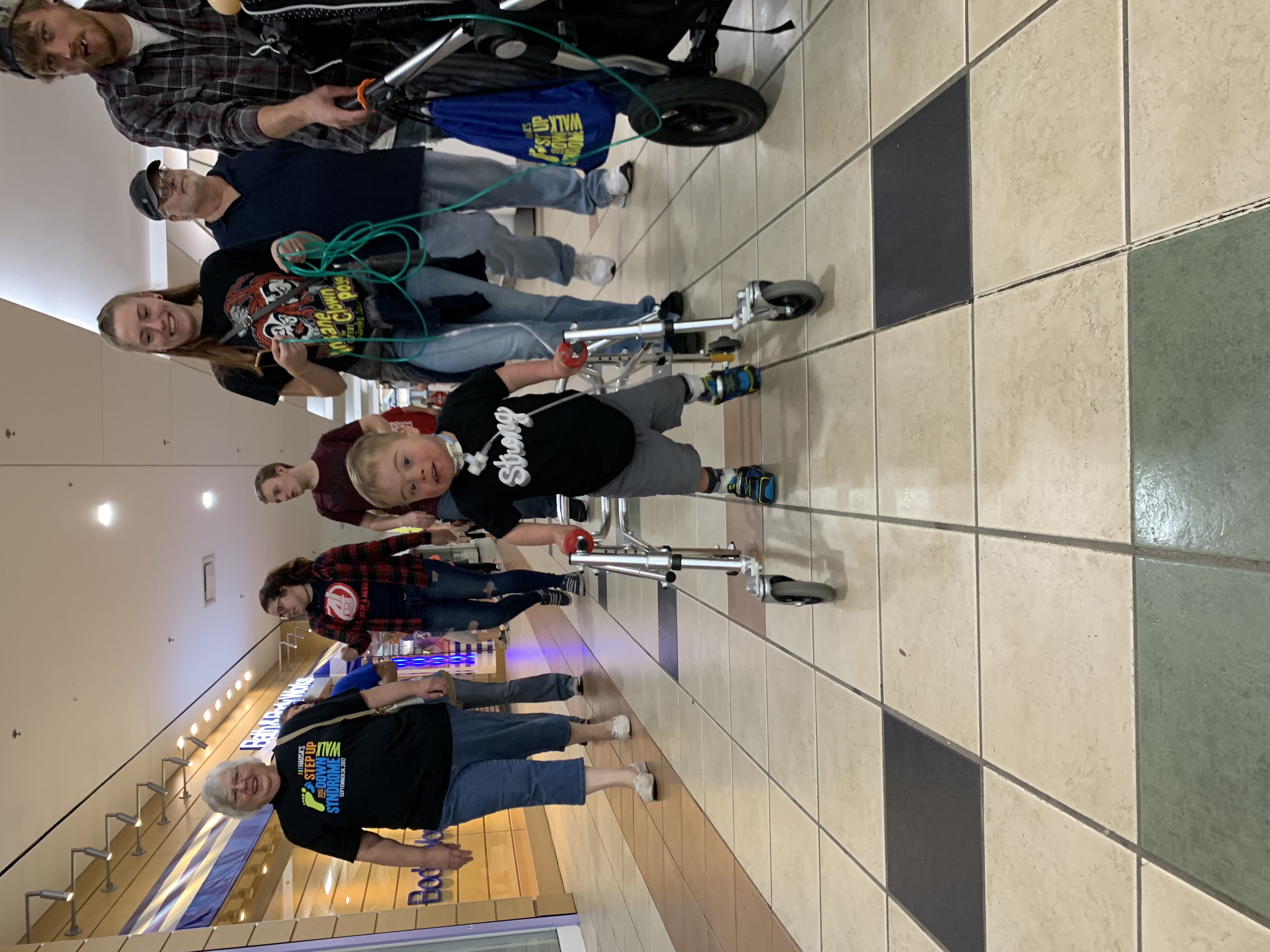 Boy with Down syndrome wearing afo and smo using a reverse walker  in  a shopping mall wearing a shirt that says strong